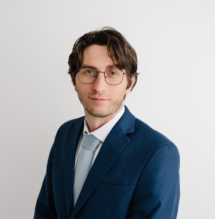Portrait of Jonathan Andrew, wearing glasses, a navy suit and a light blue tie against a plain light background.