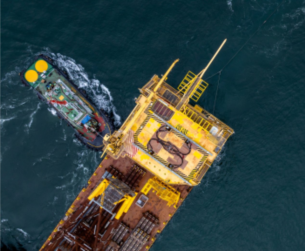 Aerial view of a yellow offshore platform and a tugboat on dark ocean water.